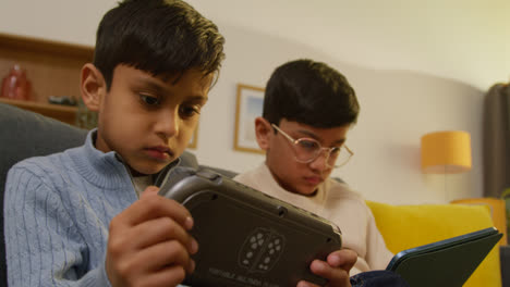 Zwei-Junge-Jungen-Sitzen-Zu-Hause-Auf-Dem-Sofa-Und-Spielen-Spiele-Oder-Streamen-Auf-Ein-Digitales-Tablet-Und-Ein-Handheld-Spielgerät-3